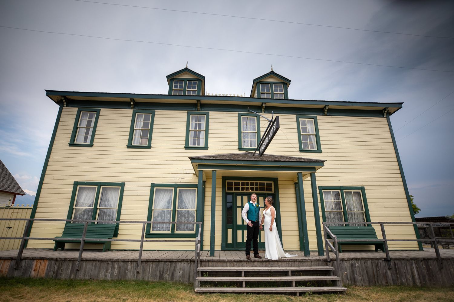 A historic yellow farmhouse with green trim and a wraparound porch sits against a cloudy sky. Fort Steele, Cranbrook, BC