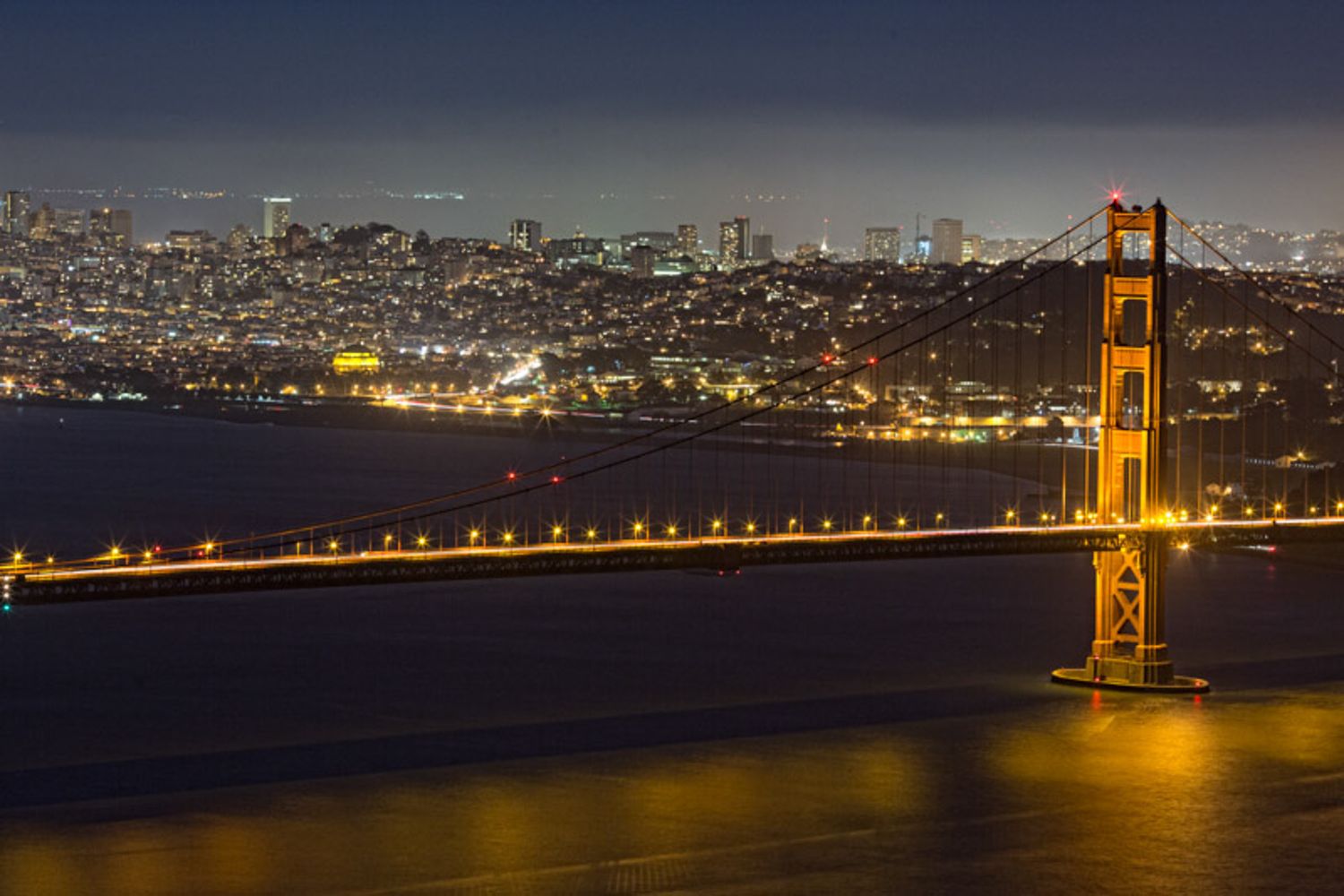 Panoramic night view of the illuminated Golden Gate Bridge spanning San Francisco Bay with city lights glowing in background.