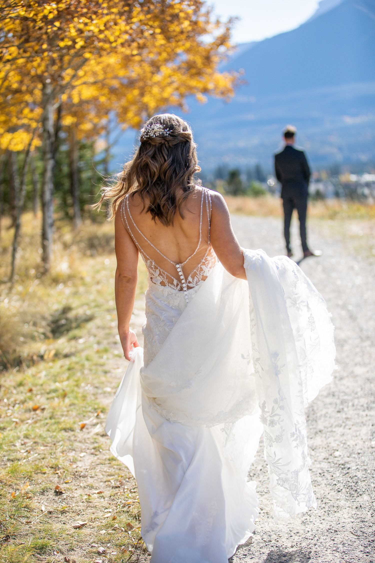 A bride in a flowing white dress walks down a mountain path surrounded by golden autumn foliage and mountain views. Benchland trail Canmore.