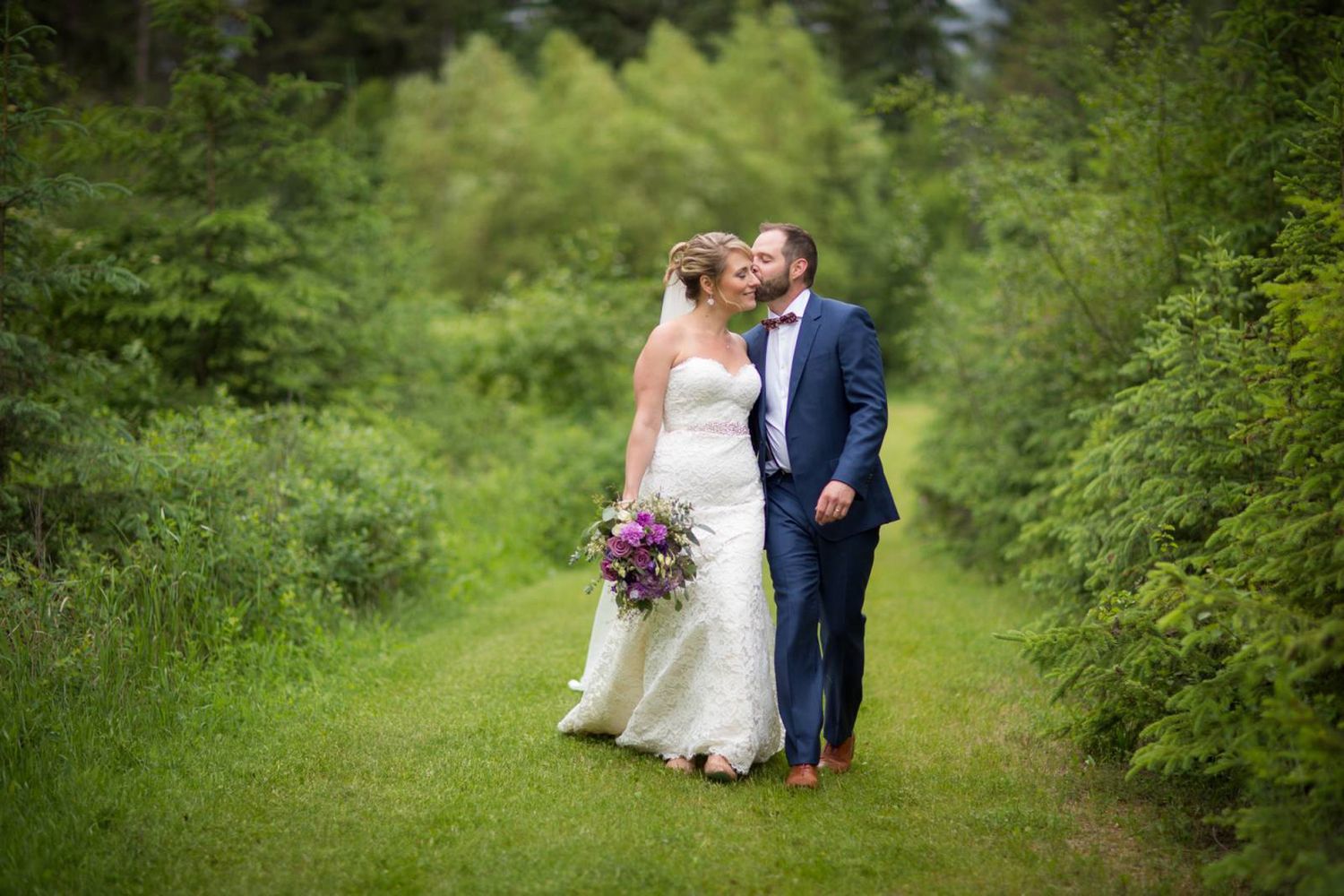 A couple in wedding attire share a romantic moment on a lush green path surrounded by foliage at the Fairmont Hot Springs, BC
