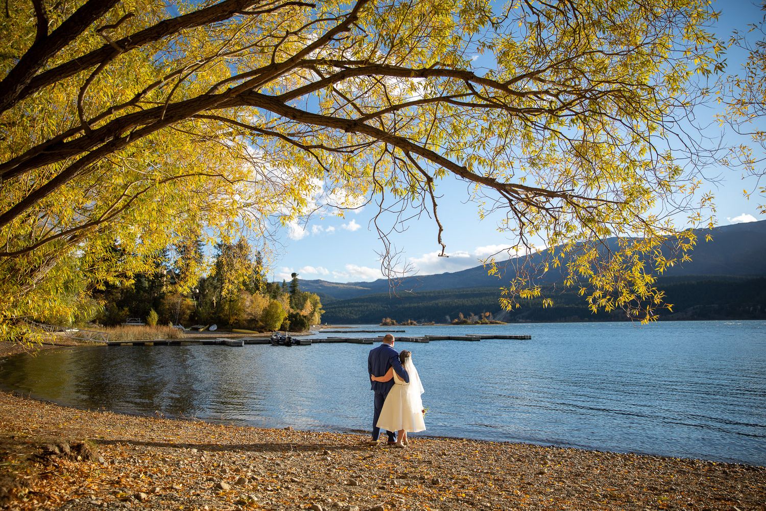 Bride and Groom stand together on the Windermere lakeside beach under golden autumn foliage with mountains in the background. Windermere, BC