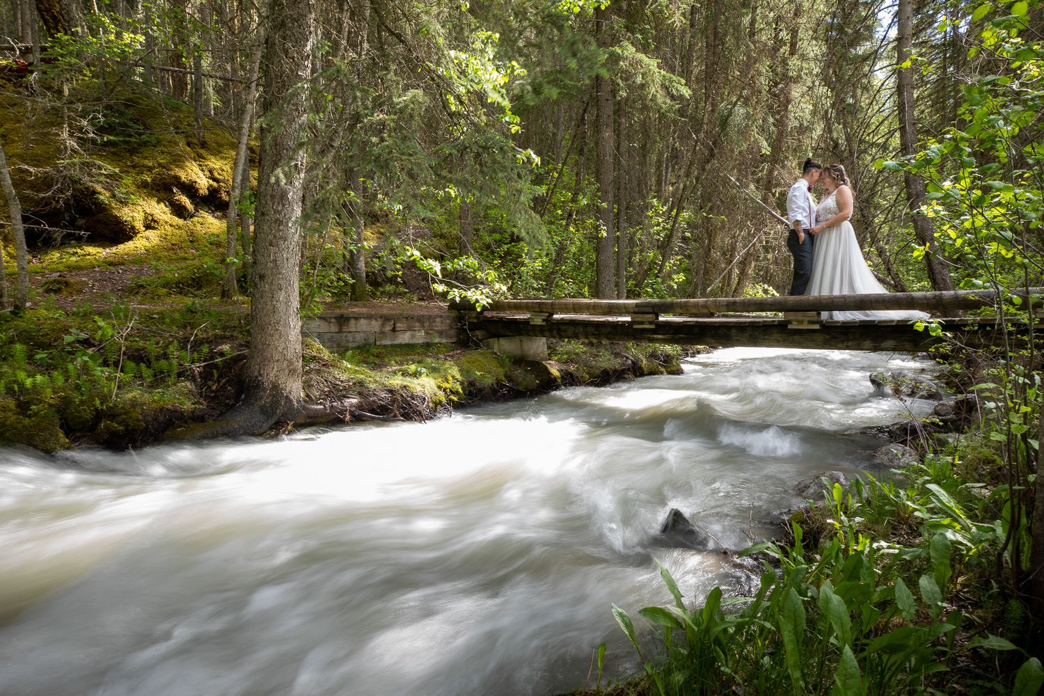 A couple stands on a wooden footbridge over rushing mountain stream in a lush forest setting. Kortney National Park in Radium, BC