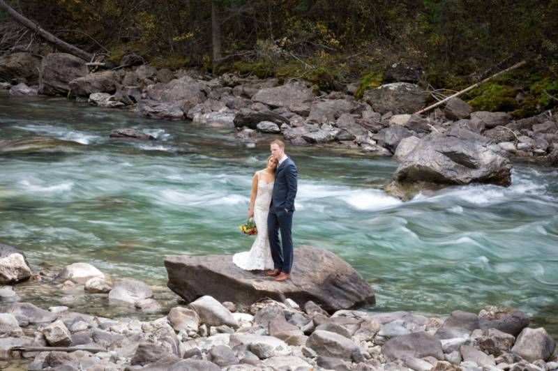 Bride and Groom stands on a large rock overlooking a rushing turquoise Toby Creek river in Panorama Resort, BC.