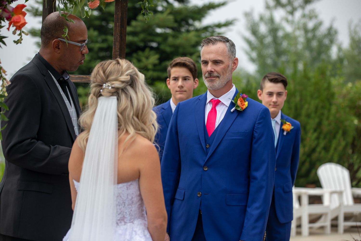 Outdoor wedding ceremony with groom in blue suit and pink tie facing bride in white dress and veil. Copperpoint resort,  Invermere.