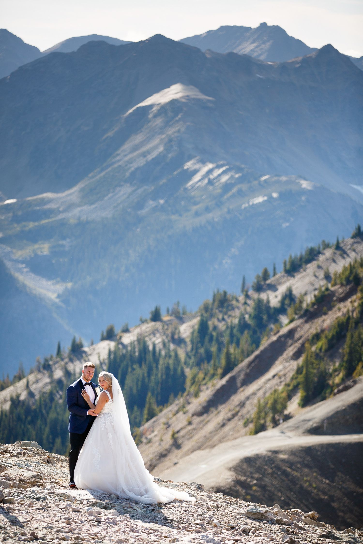 Couple stands on rugged mountain peak with dramatic snow-capped peaks and evergreen forest in the background. Kicking Horse resort, Golden,