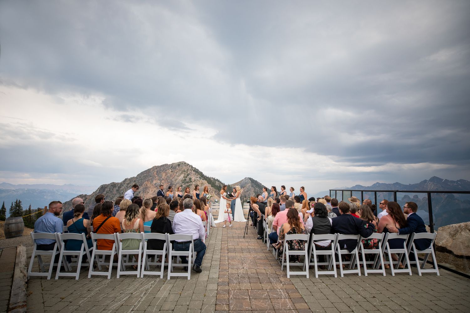 Outdoor wedding ceremony on a mountain terrace with guests seated in white chairs under a dramatic cloudy sky at Kicking Horse Resort, BC