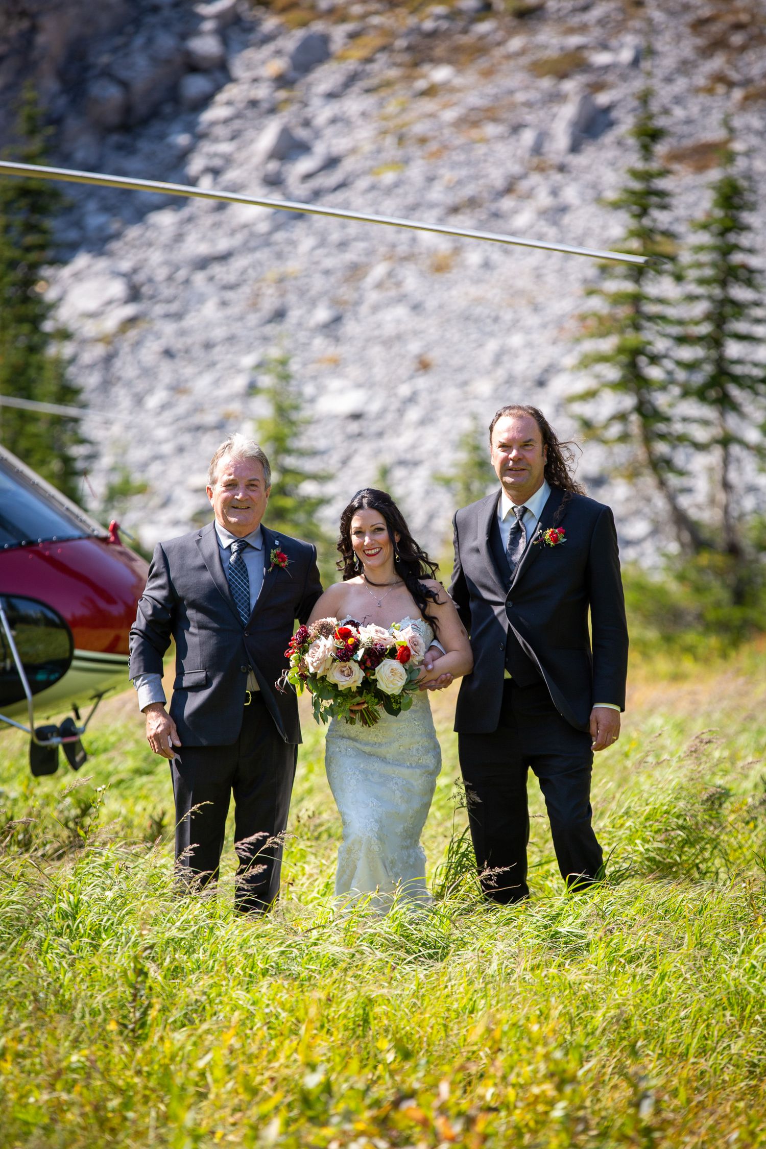 Bride with bouquet and two men in suits pose in mountain meadow with red helicopter backdrop in Kananaskis, Alberta.