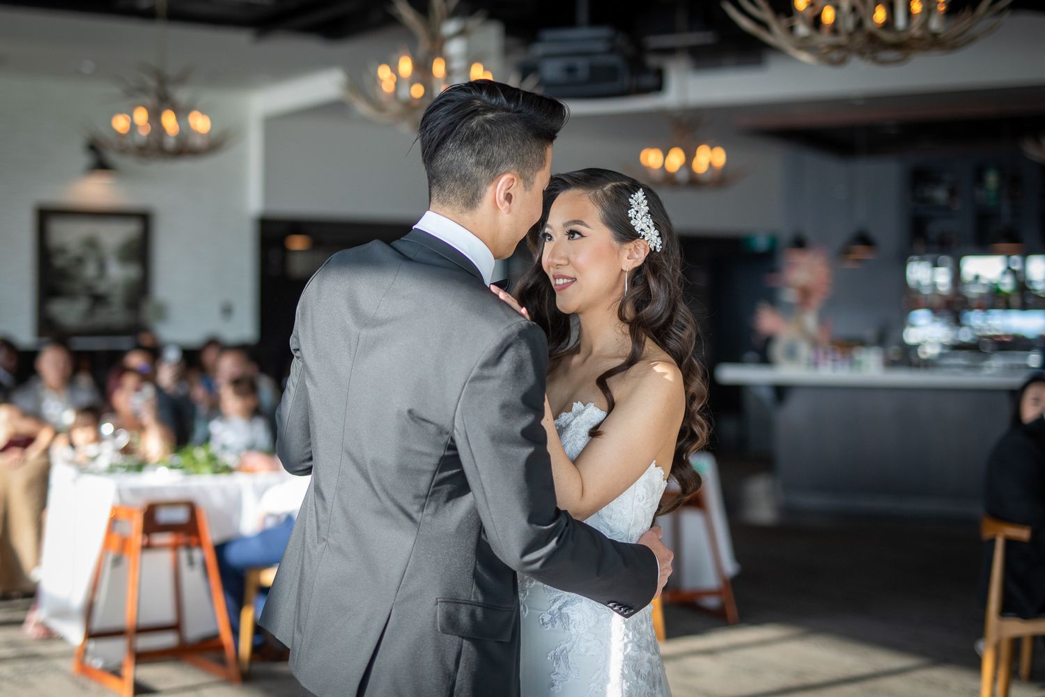 Bride and groom share their first dance at elegant wedding reception with chandeliers and guests in background.