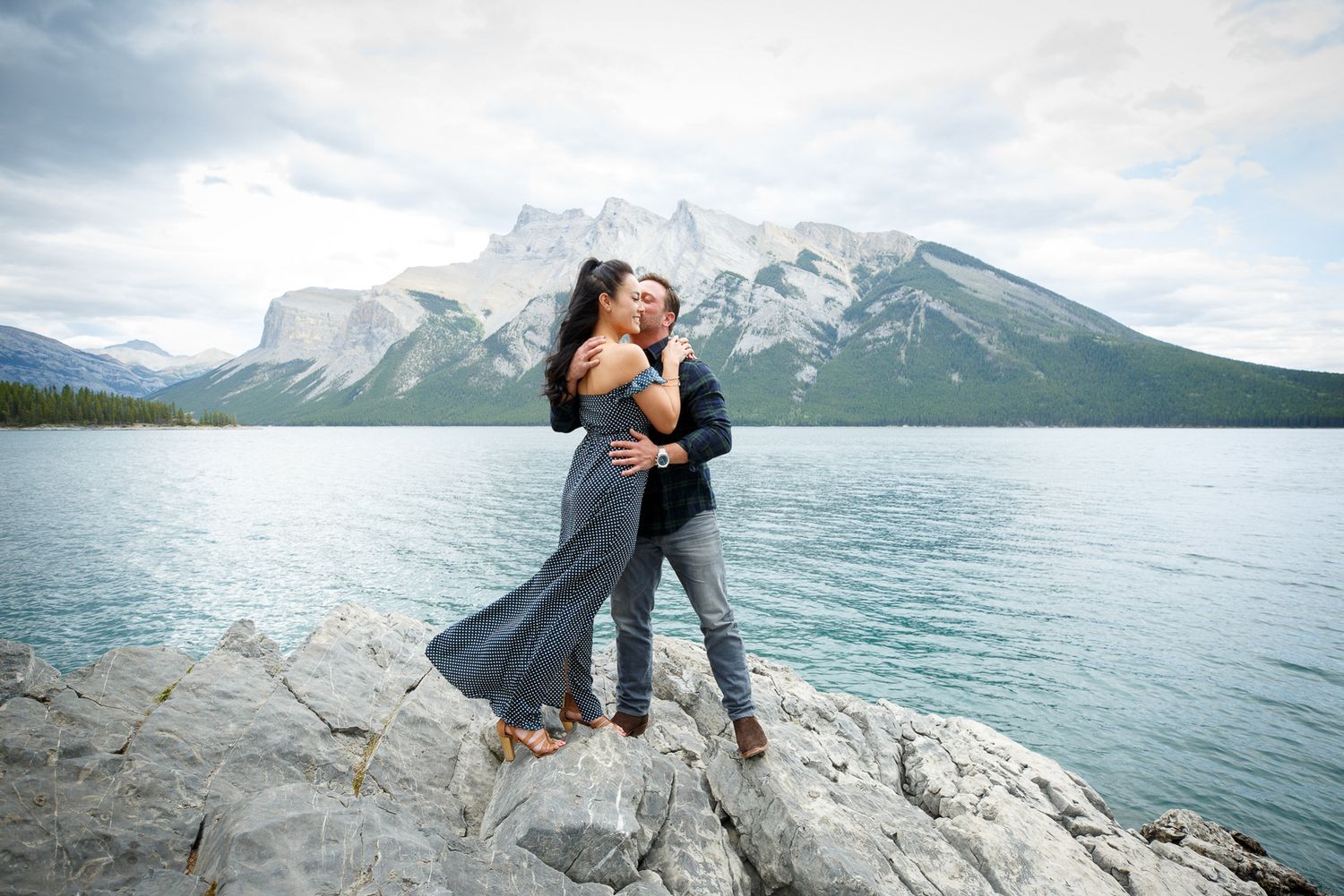 A couple embracing on a rocky shore with a scenic mountain lake and snowy peaks in the background at Lake Minnewanka in Banff.