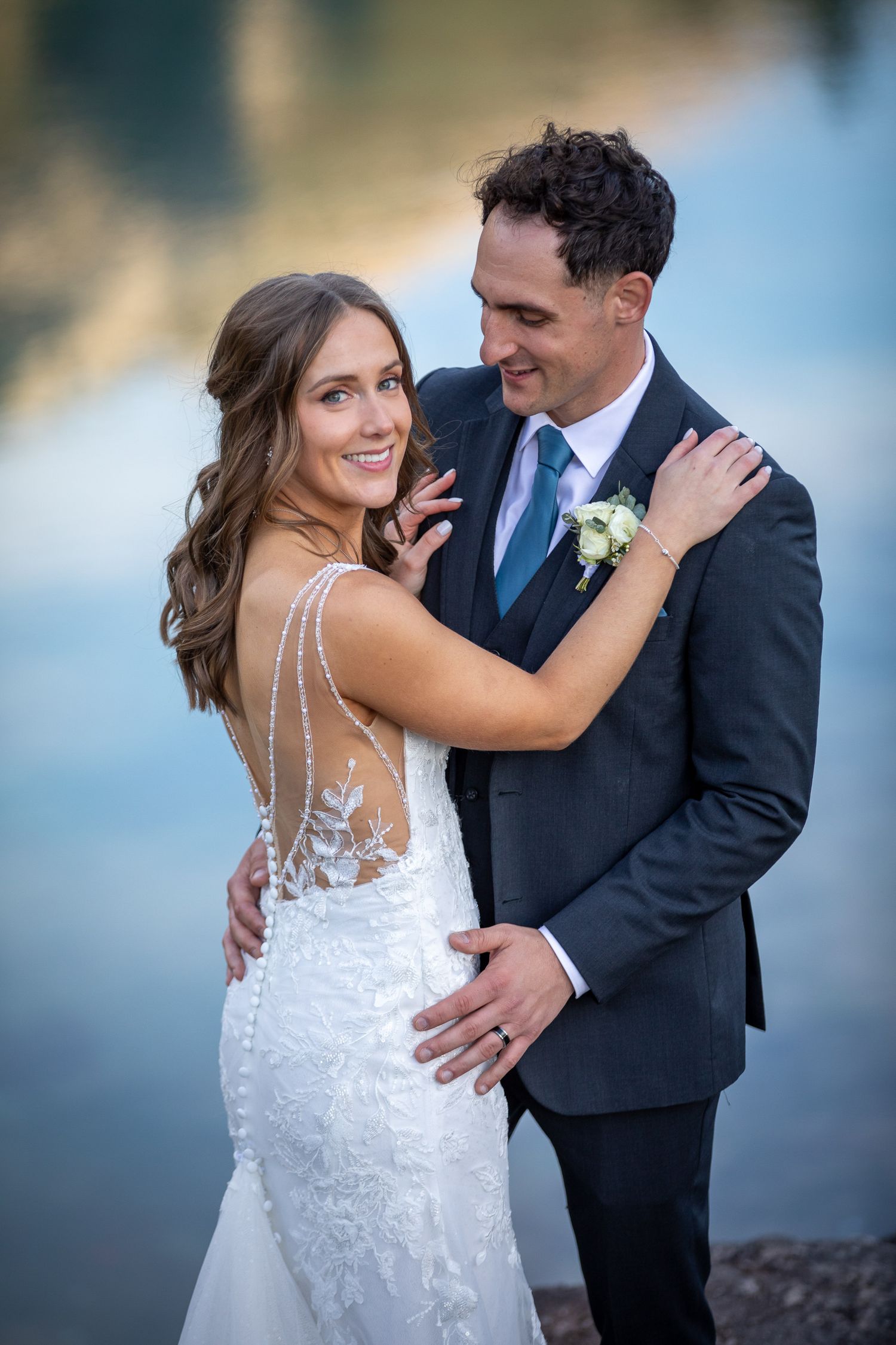 A romantic wedding couple embraces outdoors during a lakeside photoshoot in elegant white dress and navy suit at Silvertip resort in Canmore