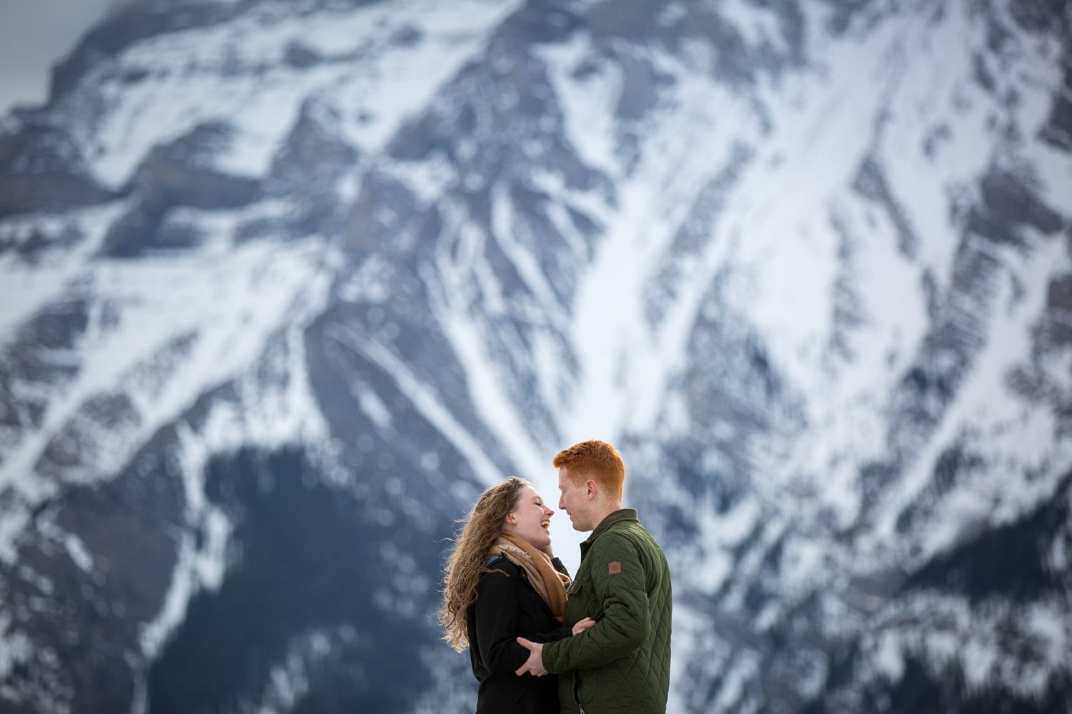 Couple embracing with snowy mountain backdrop in winter engagement photo session at Lake Minnewanka in Banff National Park.