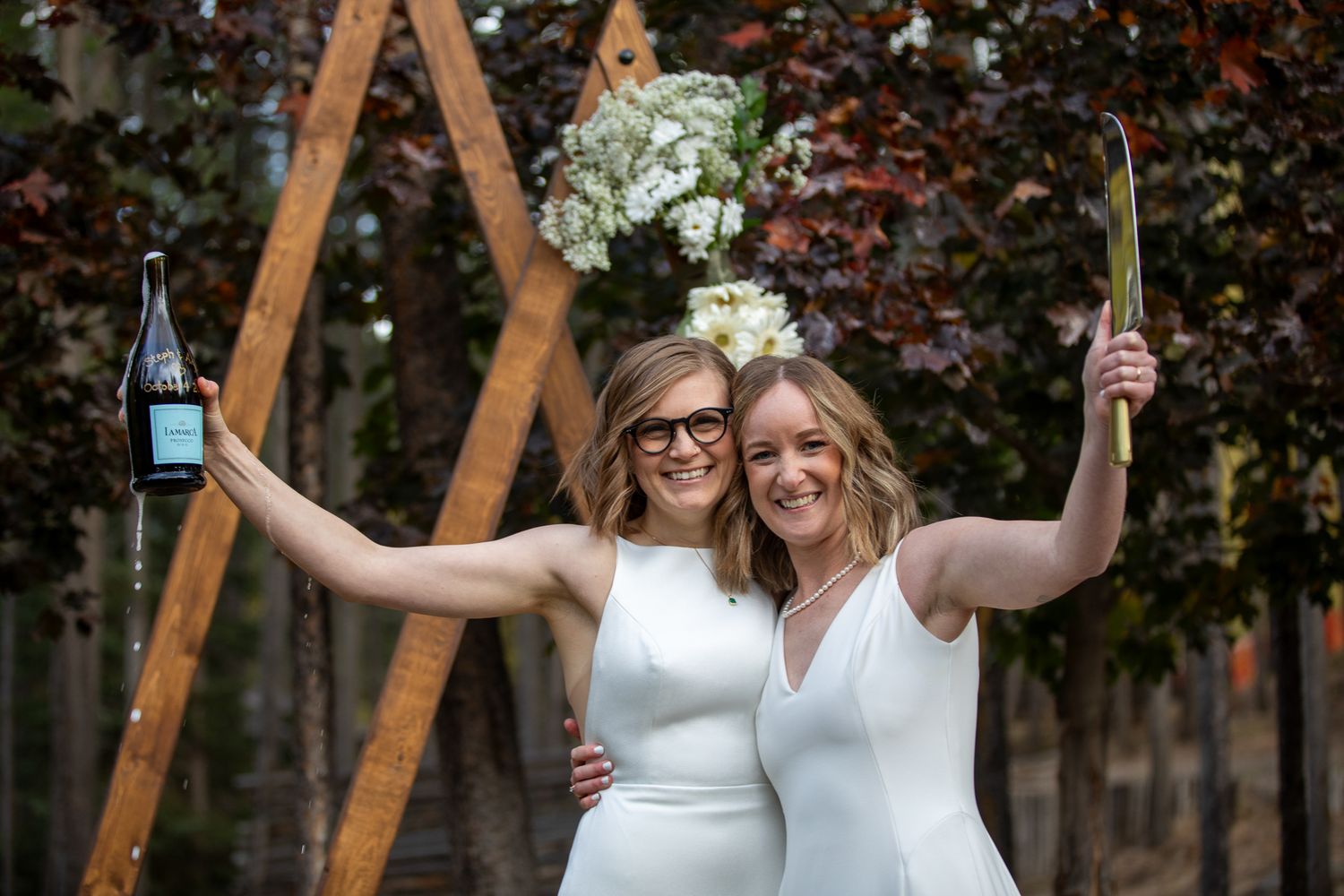 A LGBTQ couple in white dresses celebrate their wedding day with raised arms under a wooden arch in an outdoor garden setting. Panorama, BC