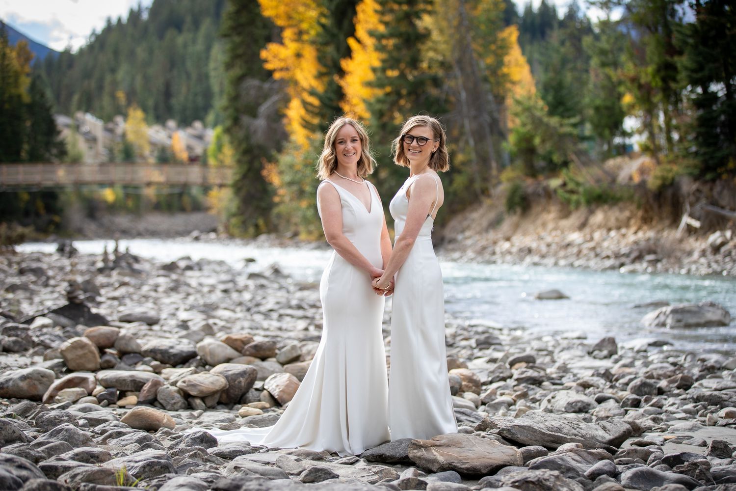A LGBTQ couple in white dresses stand together on a rocky riverbank surrounded by autumn trees and mountains. Earl Grey Lodge Panorama BC