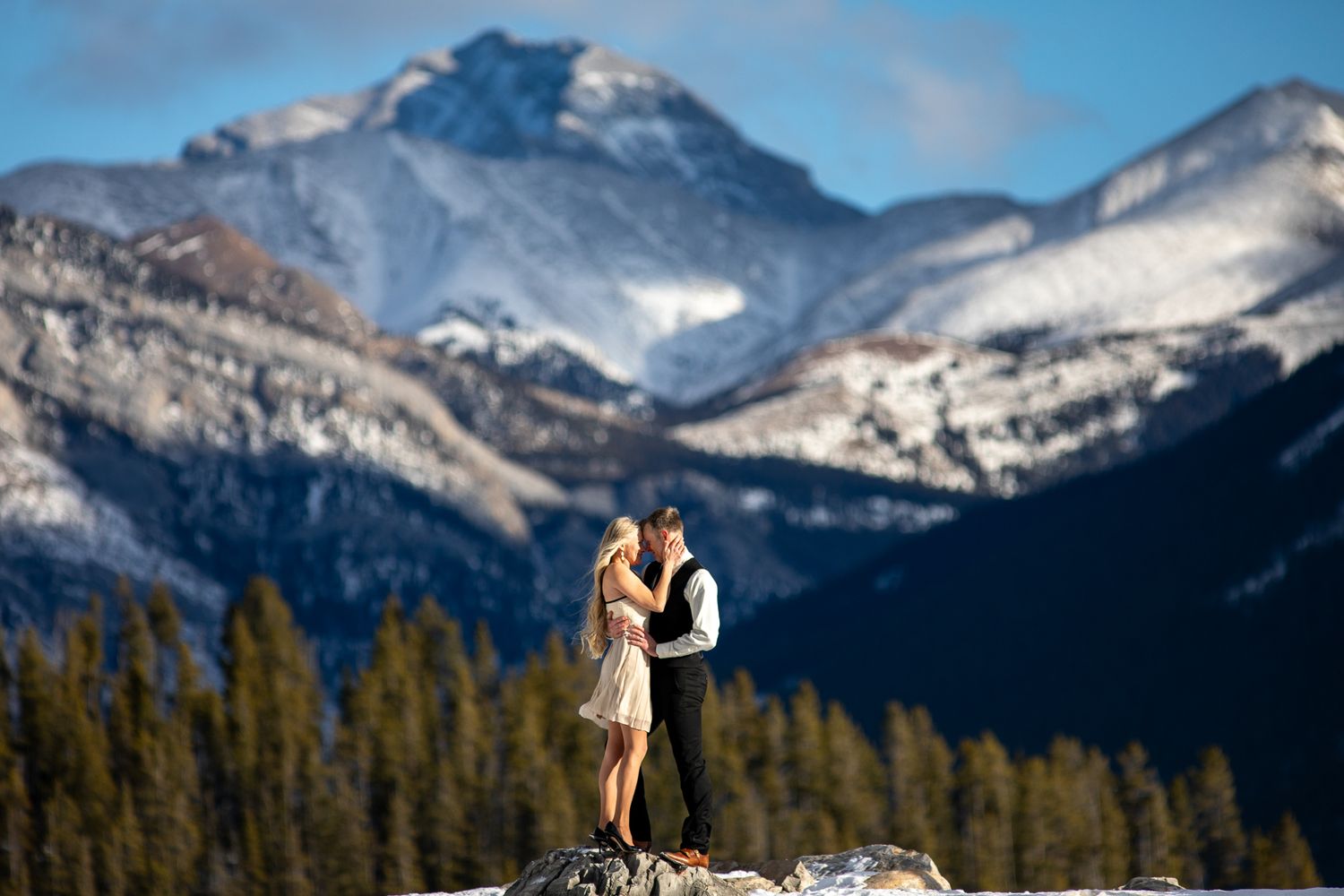 A couple embraces on a rocky outcrop with snow-capped Rocky Mountains and evergreen forest in the background at Lake Minnewanka in Banff.