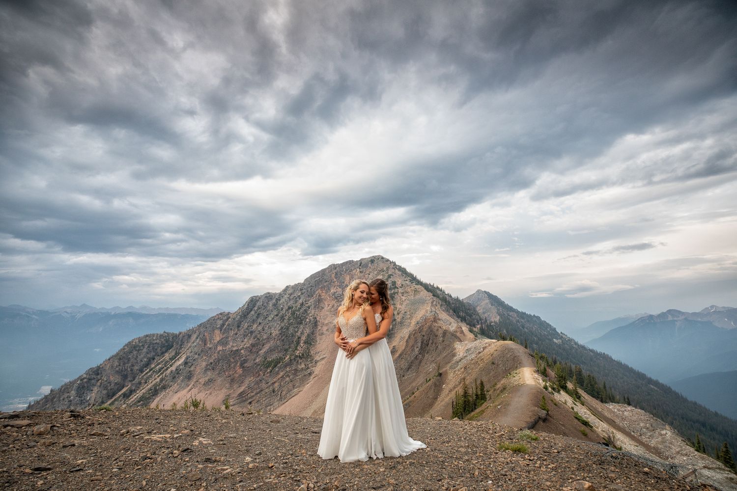 Two brides embraces on a mountain summit with dramatic cloudy skies and rugged peaks in the background at Kicking Horse resort in Golden, BC