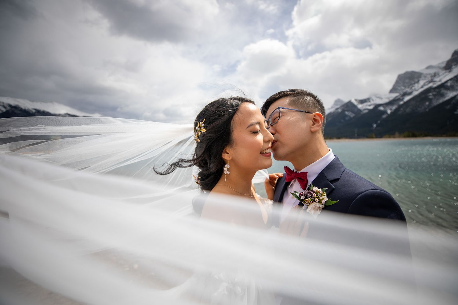 A romantic wedding couple embraces against a dramatic mountain landscape with a veil in the wind by Bow river in Canmore, AB.