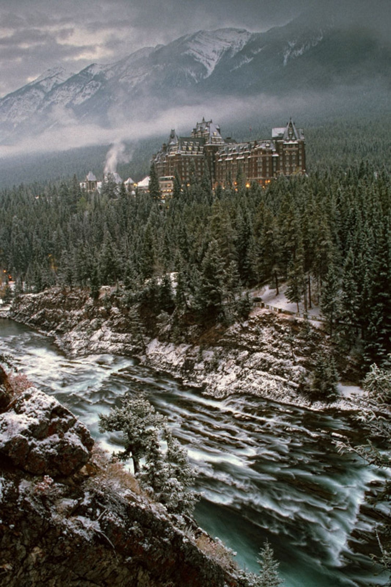 The historic Banff Springs Hotel perches dramatically above a rushing river amid snowy mountains and evergreen forests in Canada.