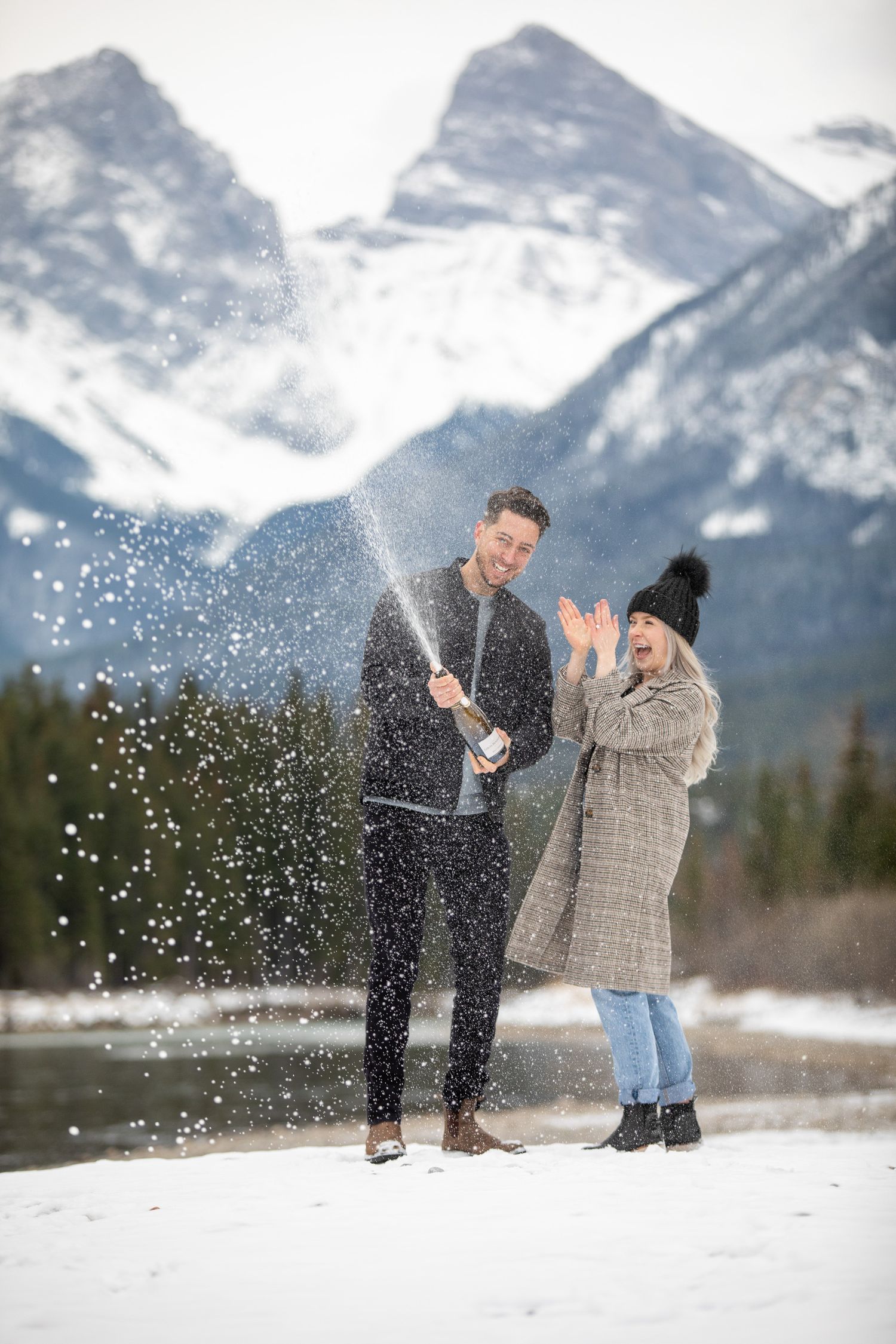 A couple celebrates with champagne in the snow against a backdrop of majestic Three Sisters mountains by Bow river in Canmore, AB.