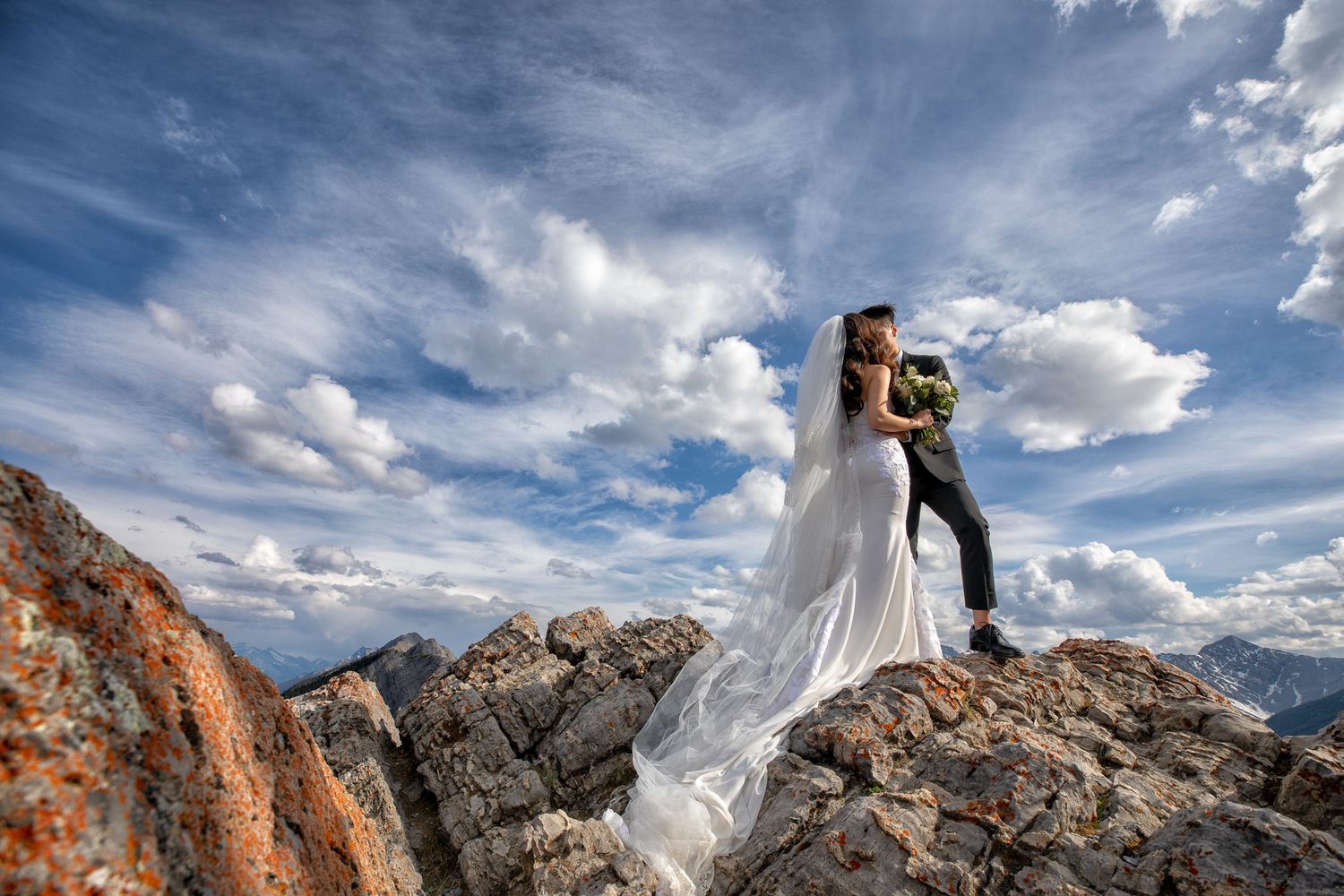 A couple poses on rocky mountain peaks against dramatic cloudy skies during their wedding ceremony on Sulphur mountain in Banff  Alberta.