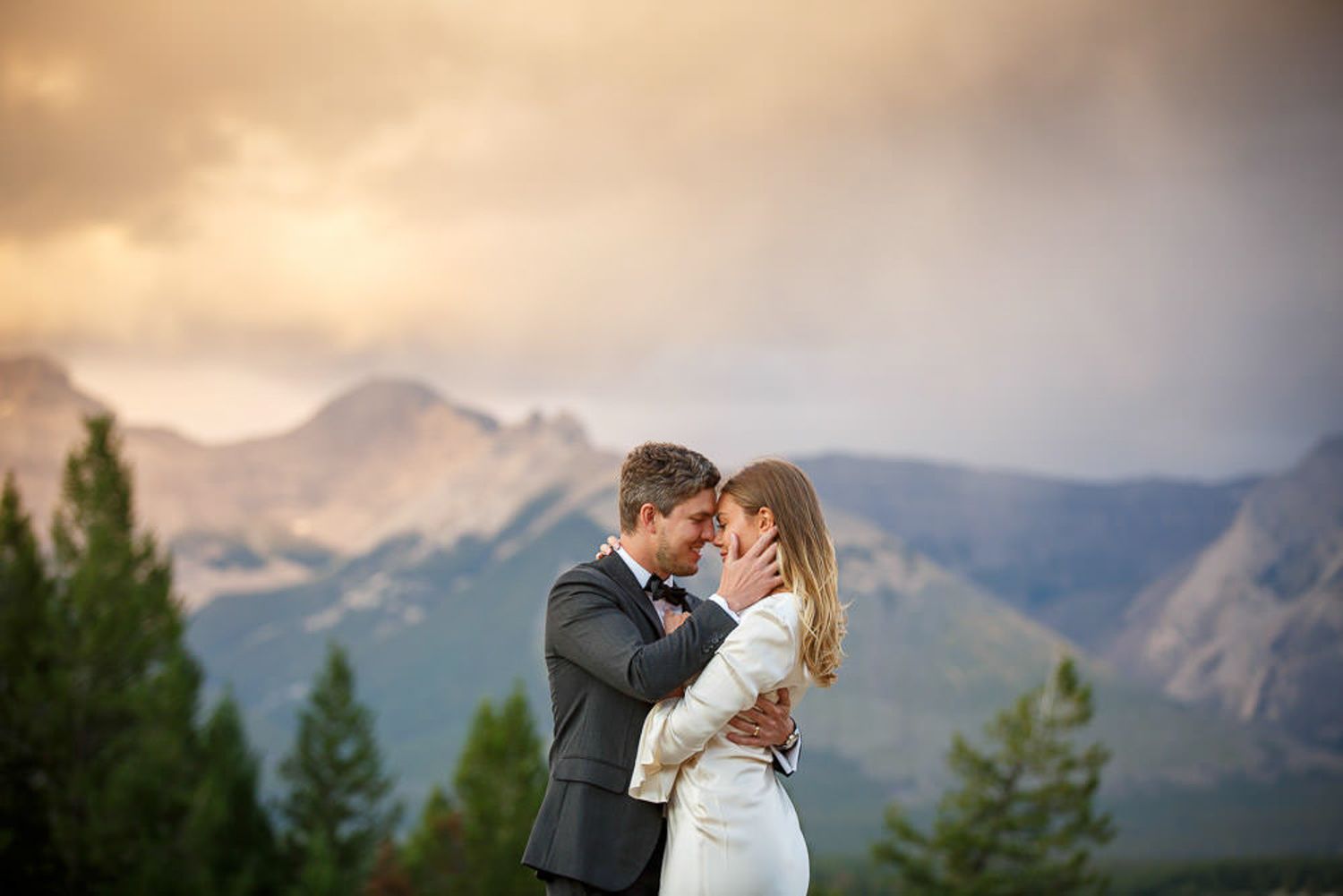A couple shares a romantic kiss against a backdrop of majestic mountains during golden hour in Banff, AB.