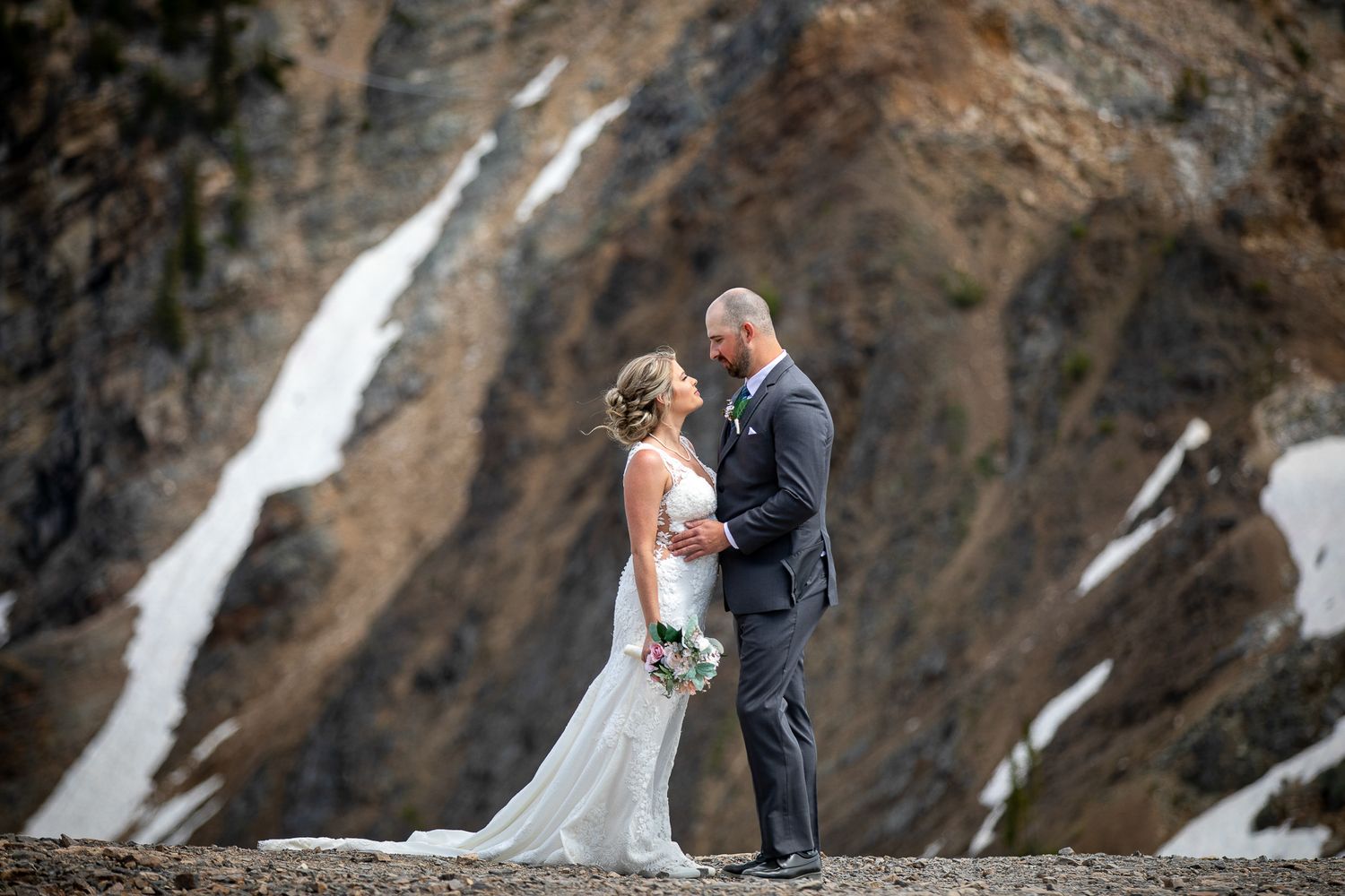 A bride and groom shares an intimate moment against a dramatic mountain backdrop with snow patches. Kicking Horse Resort, Golden, BC.