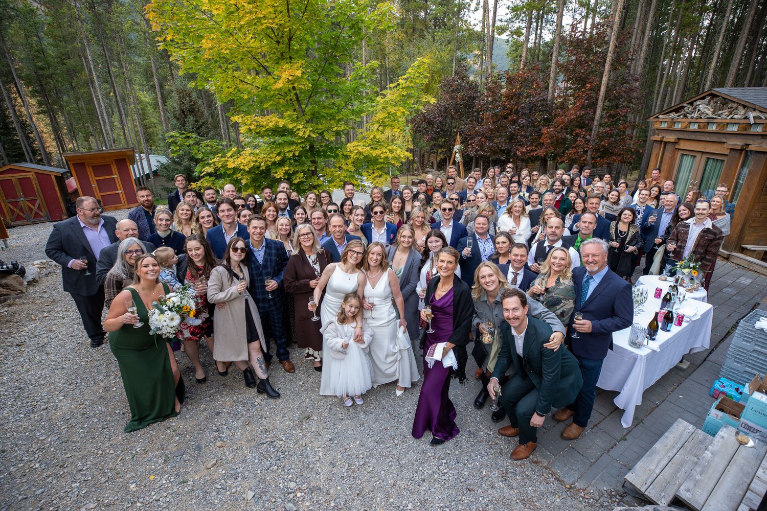 Large group of wedding guests gathered outdoors in front of a rustic venue for a panoramic photo. Earl Grey Lodge Panorama BC