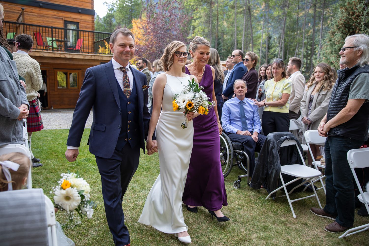 A couple walks down the aisle during an outdoor wedding ceremony while guests look on from white folding chairs. Earl Grey Lodge Panorama BC