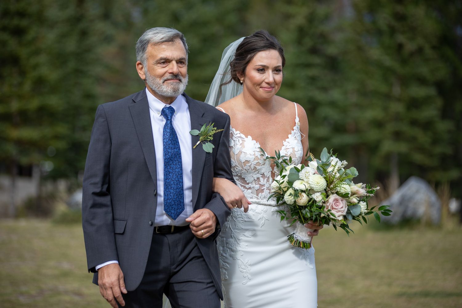 A bride in a white lace wedding dress walks down a wooded path with a bouquet of white flowers. Banff, AB