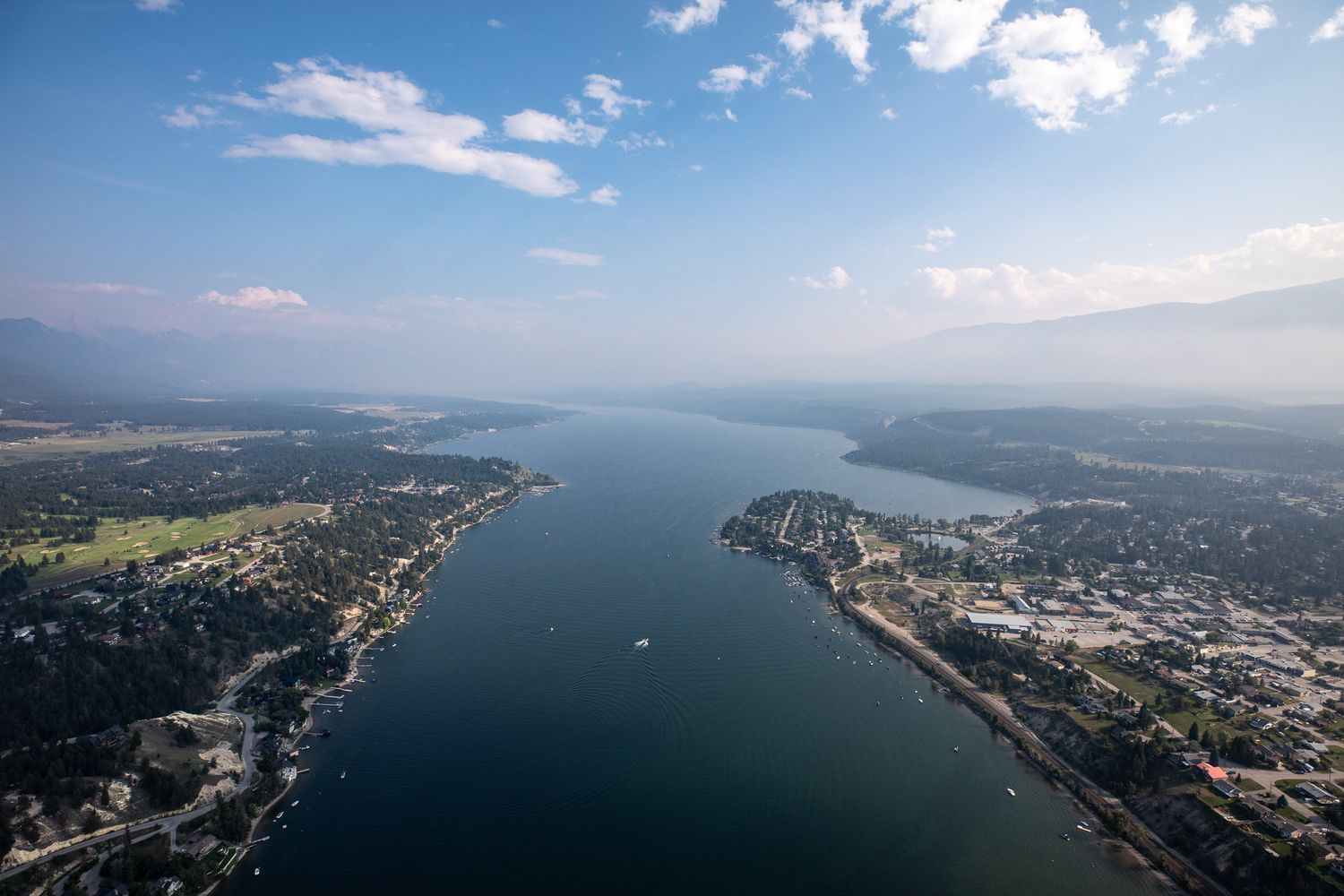 Aerial view of a scenic Windermere lake town with blue waters, lush greenery, and mountains under a partly cloudy sky over Invermere, BC.