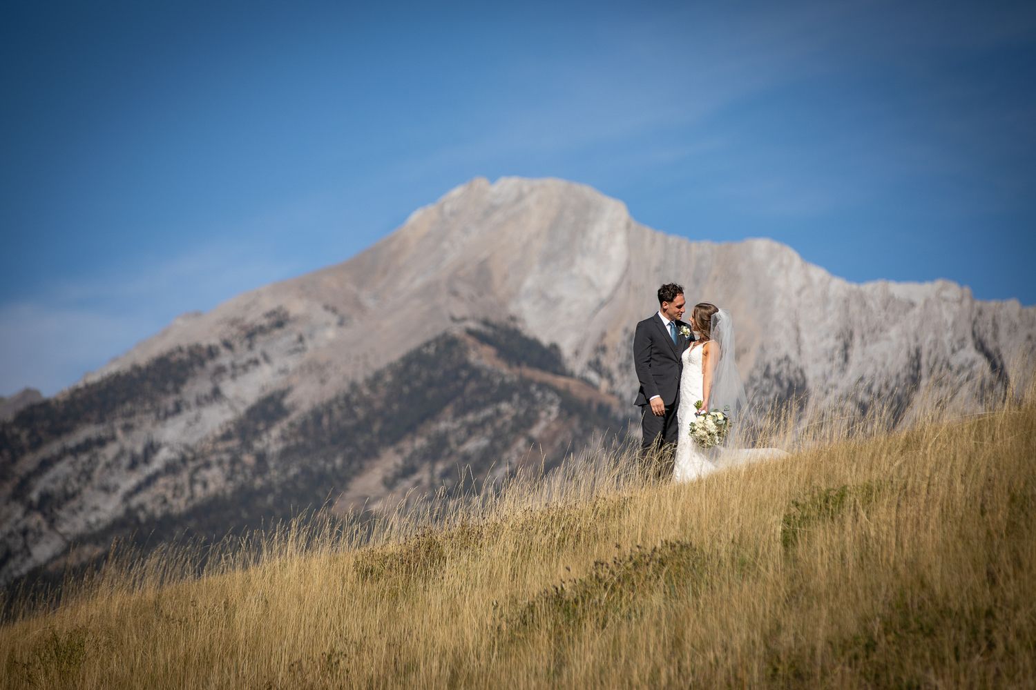 A couple embraces on a grassy hillside with majestic mountain peaks rising against a blue sky in the background at Quarry lake in Canmore.