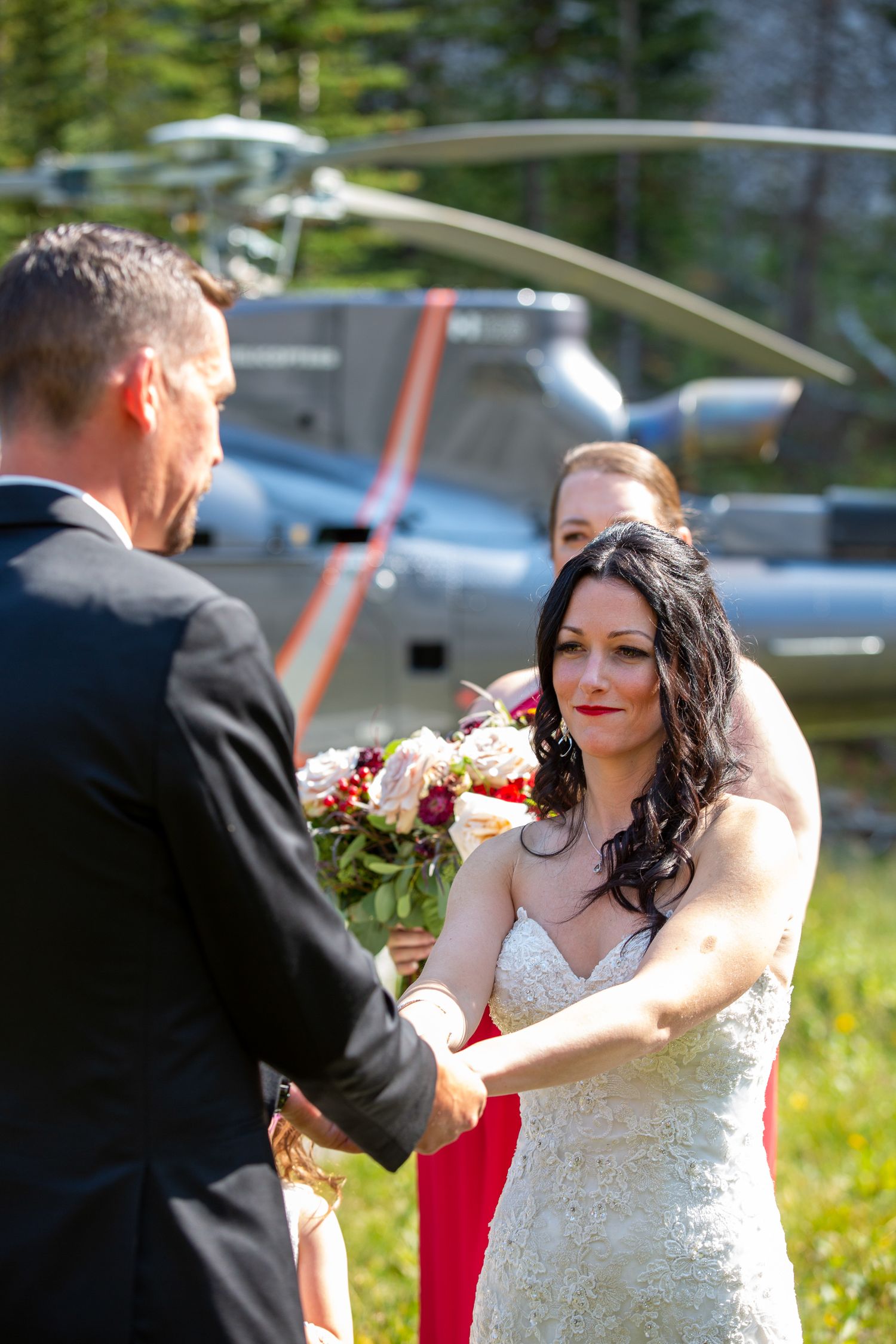 Bride in lace gown holds bouquet while exchanging vows with groom, helicopter visible in background outdoors in Kananaskis, Alberta.