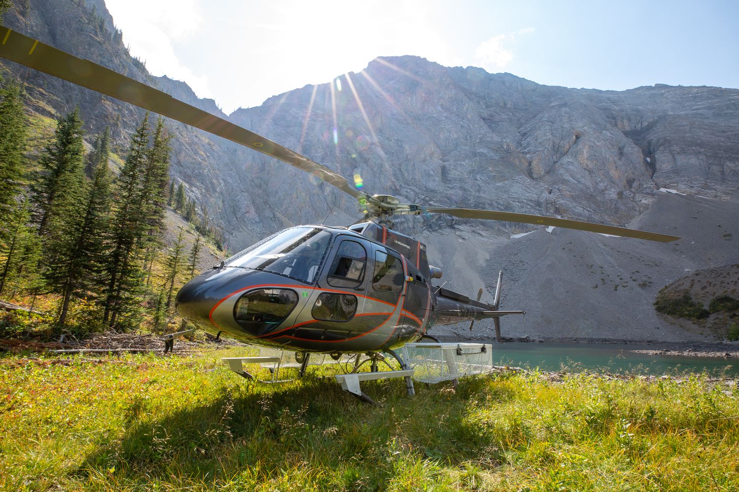 Helicopter landed in alpine meadow with mountain lake and sunburst, surrounded by rocky peaks and autumn foliage in Kananaskis, Alberta.