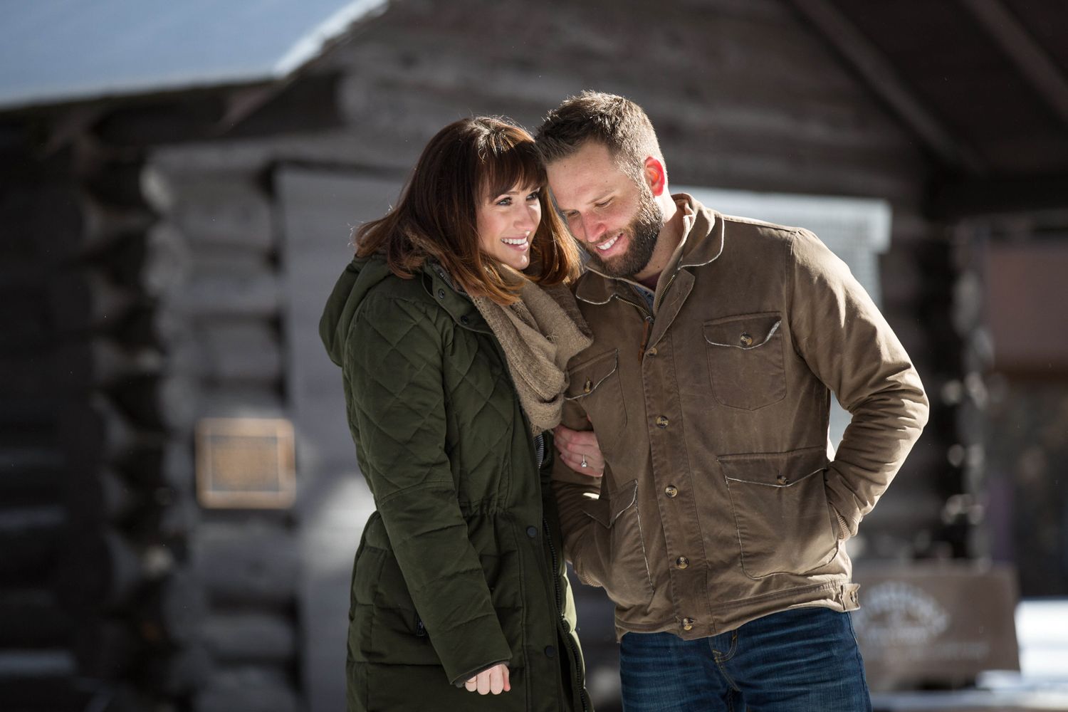 A couple in casual winter clothing embrace intimately against a log cabin background in Banff, AB.
