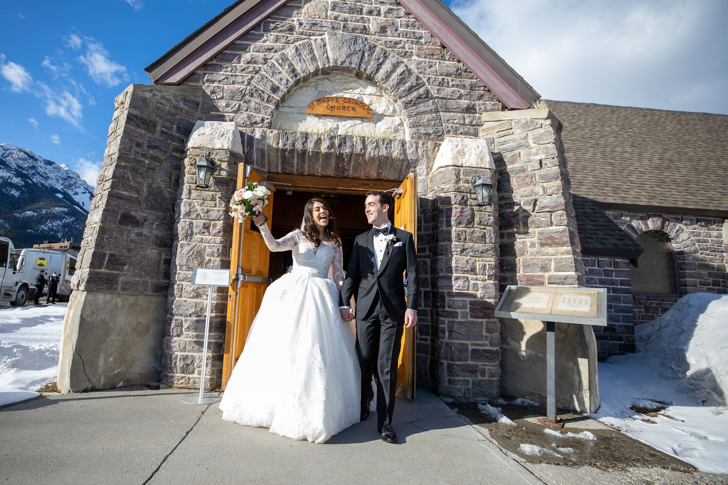 A newly married couple stands outside a stone church building in a snowy winter landscape in Banff, AB.