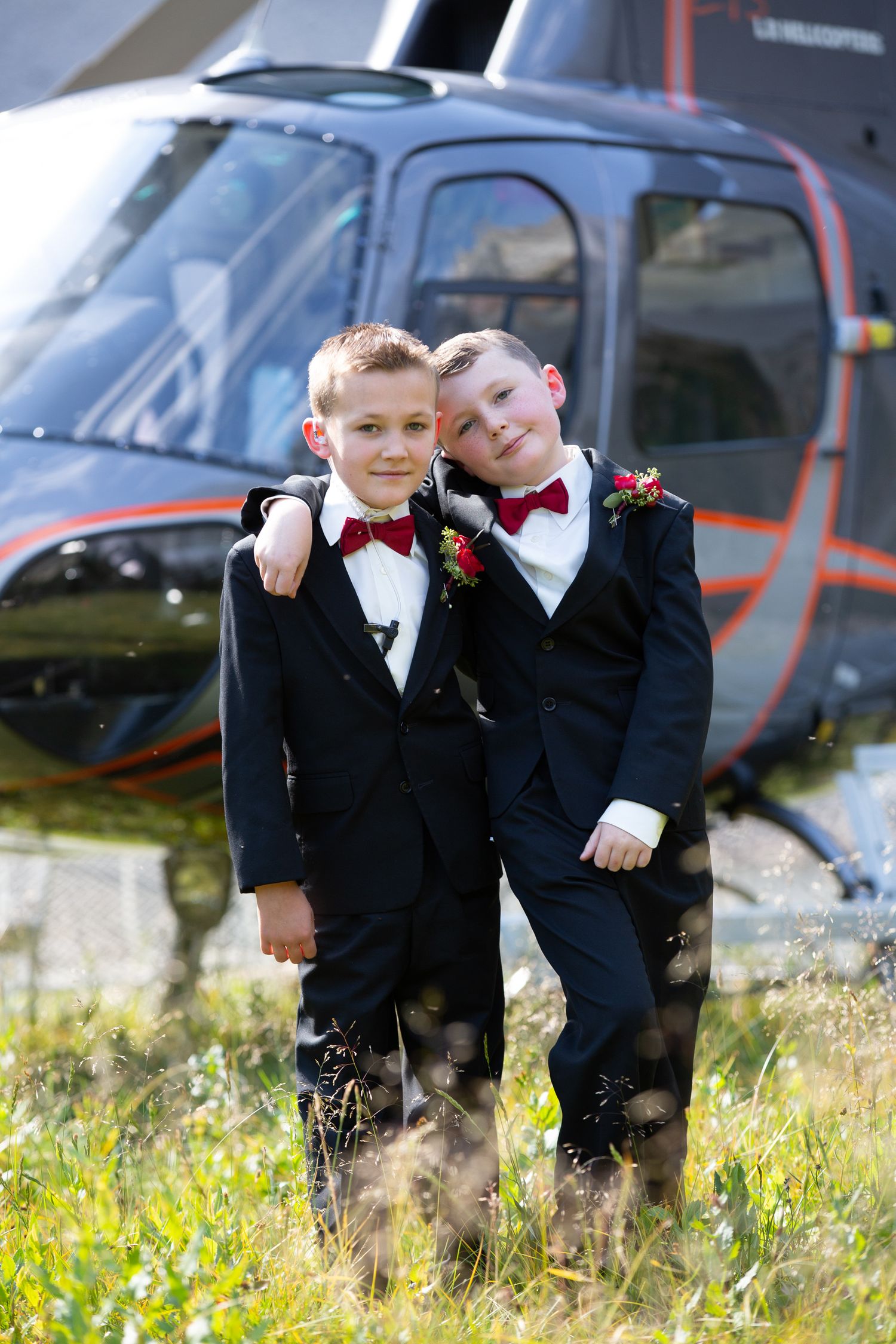 Two boys in black tuxedos with red bow ties pose together in front of a helicopter outdoors.