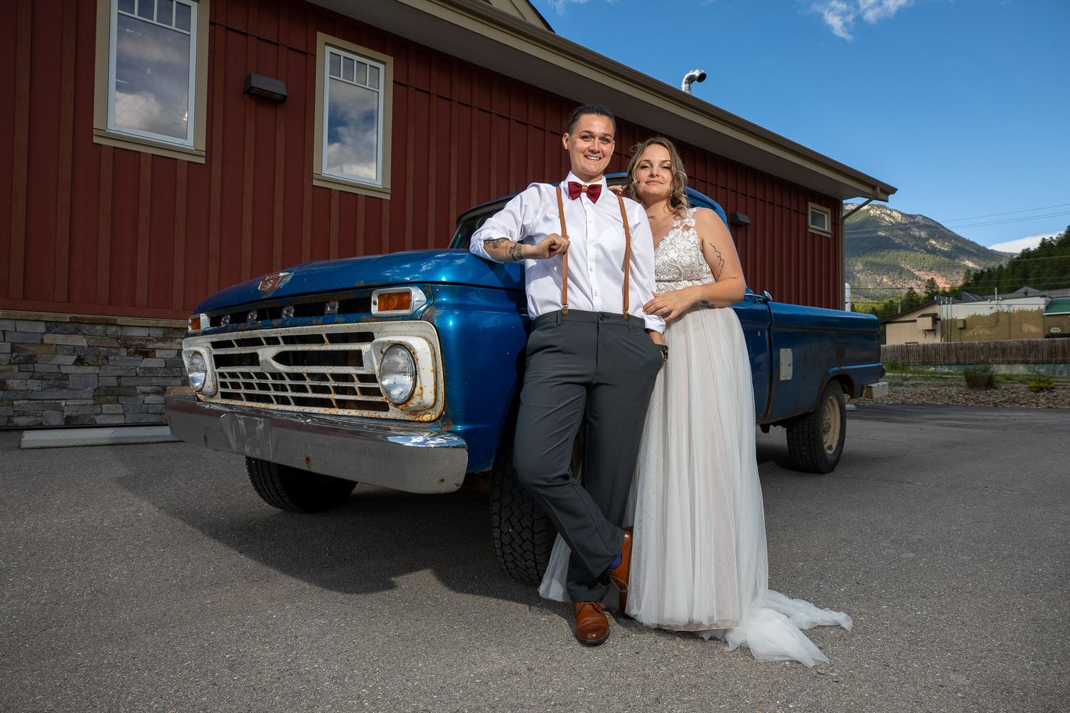 A couple poses together outdoors near a red barn and vintage car on a sunny day. Radium-Invermere, British Columbia.