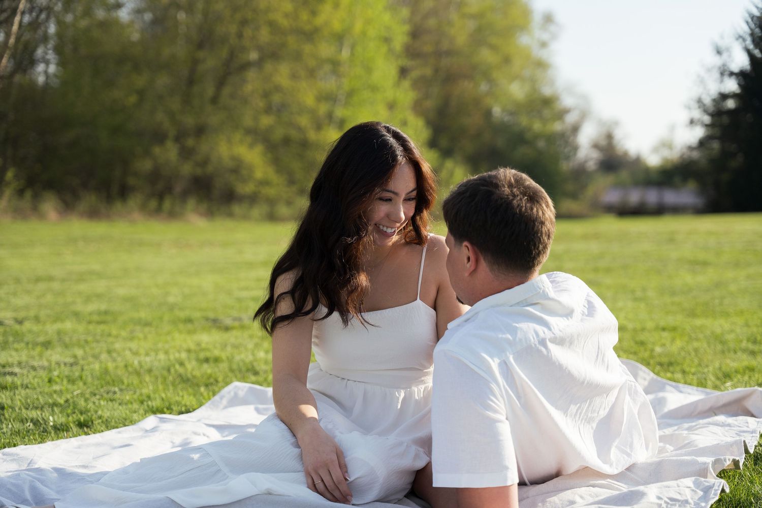 A couple shares a romantic moment on a white blanket in a sunny green park, the woman in a white dress smiling warmly.