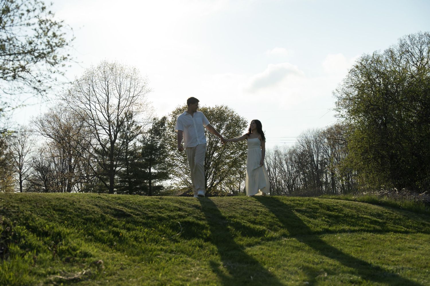A couple in white outfits holds hands while walking on a grassy hill surrounded by trees under a bright sky.