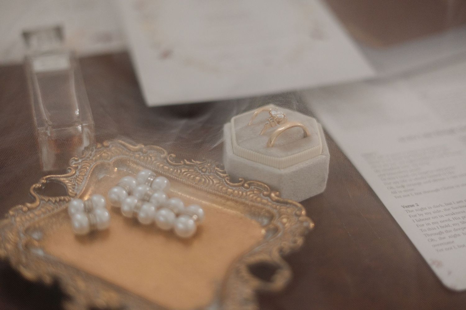 Pearl earrings on an ornate gold vanity tray beside a ring in a white hexagonal box and a glass perfume bottle on a wooden table.