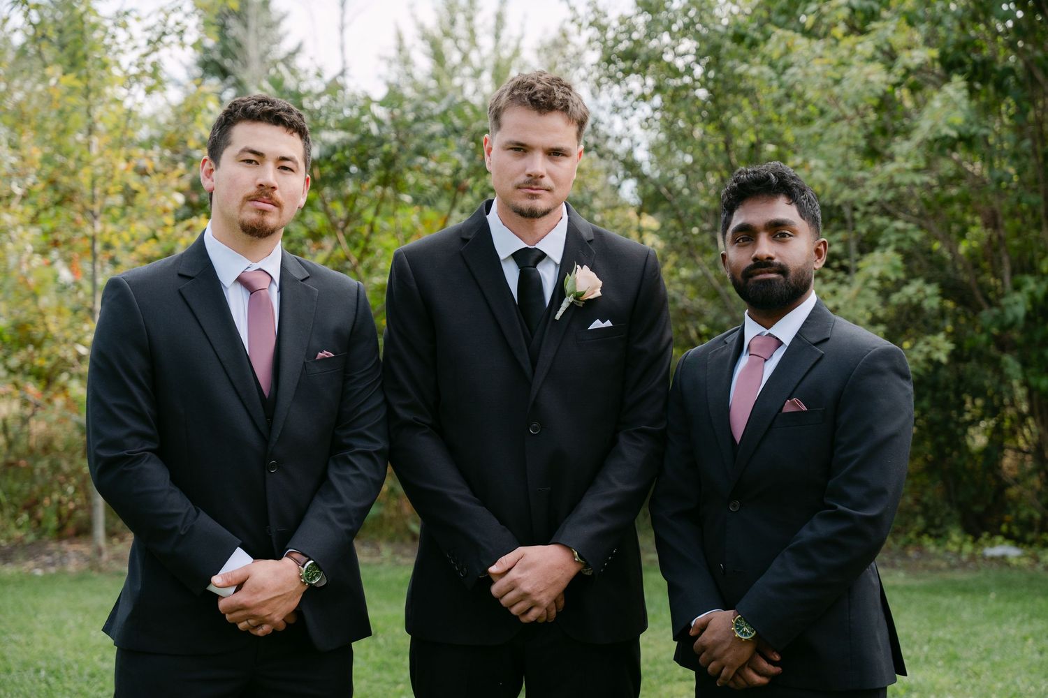 Three groomsmen in dark suits and ties stand side by side outdoors on a green lawn, one wearing a boutonniere, with trees in the background.