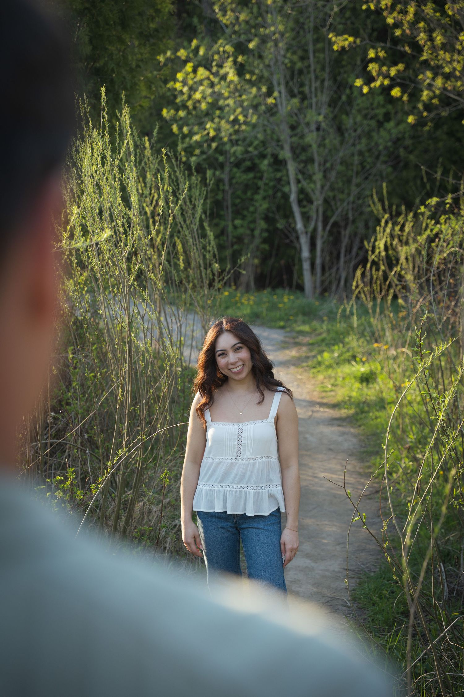 Young woman in white top and jeans smiling on a nature trail, with lush green trees in the background.