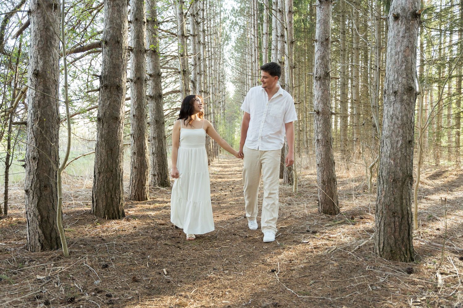 Couple holding hands walking through a pine forest, woman in white dress, man in white shirt and khaki pants.