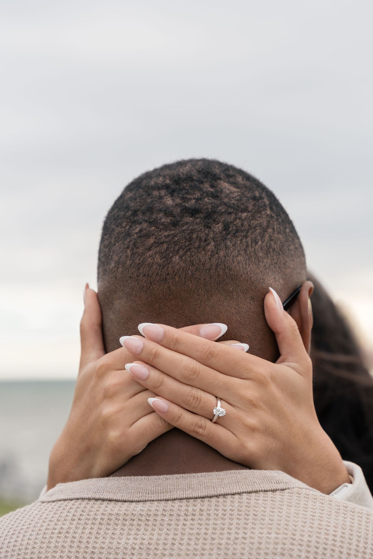 Close-up of hands with engagement ring wrapped around someone's head against a cloudy sky.