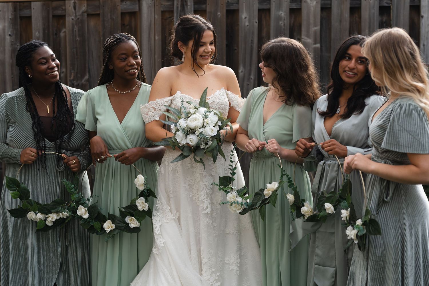 Bridesmaids in sage green dresses hold white bouquets while standing with the bride.