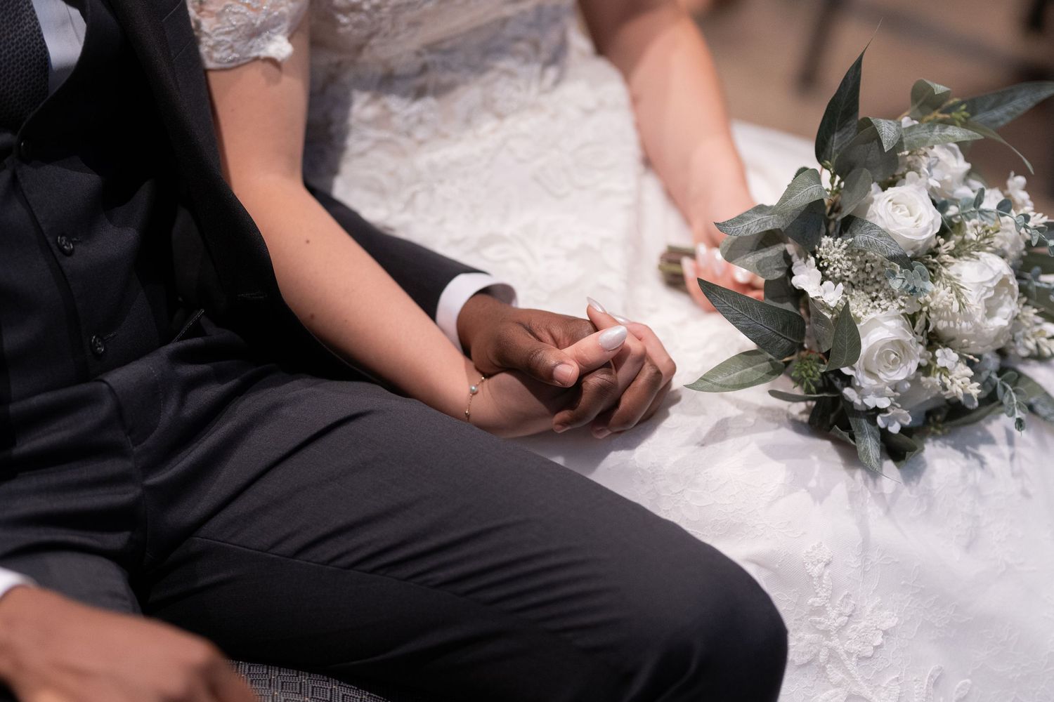 A close-up of hands holding together with a white bridal bouquet laying nearby.