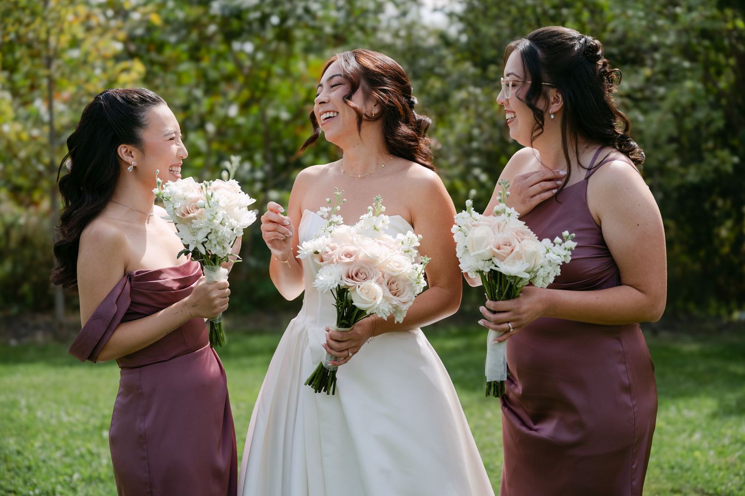 A bride in a white gown laughs with two bridesmaids in mauve dresses as they hold white and blush bouquets on a sunny lawn.