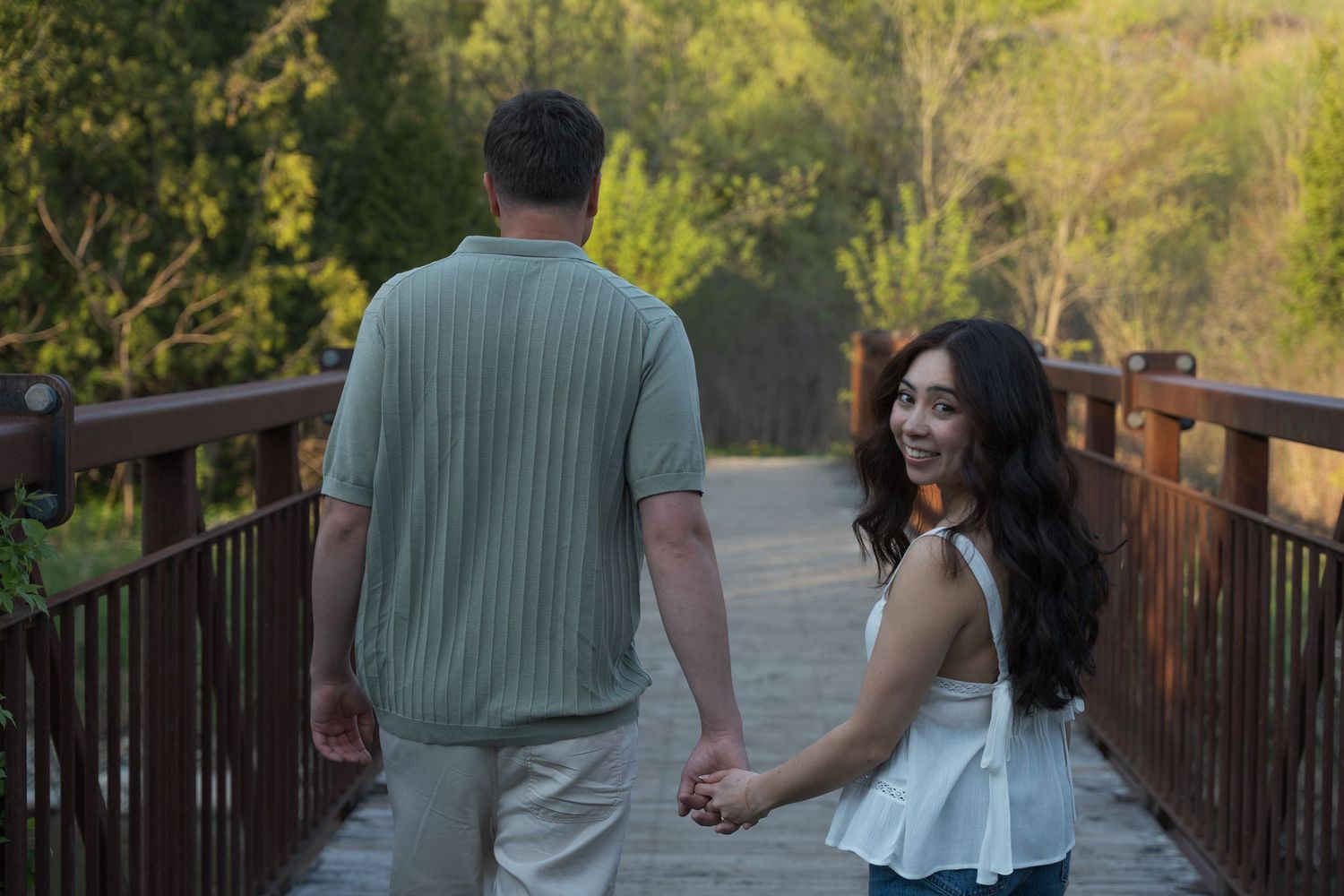 Couple holding hands on a wooden bridge, woman in white top looking back, man in sage green shirt.