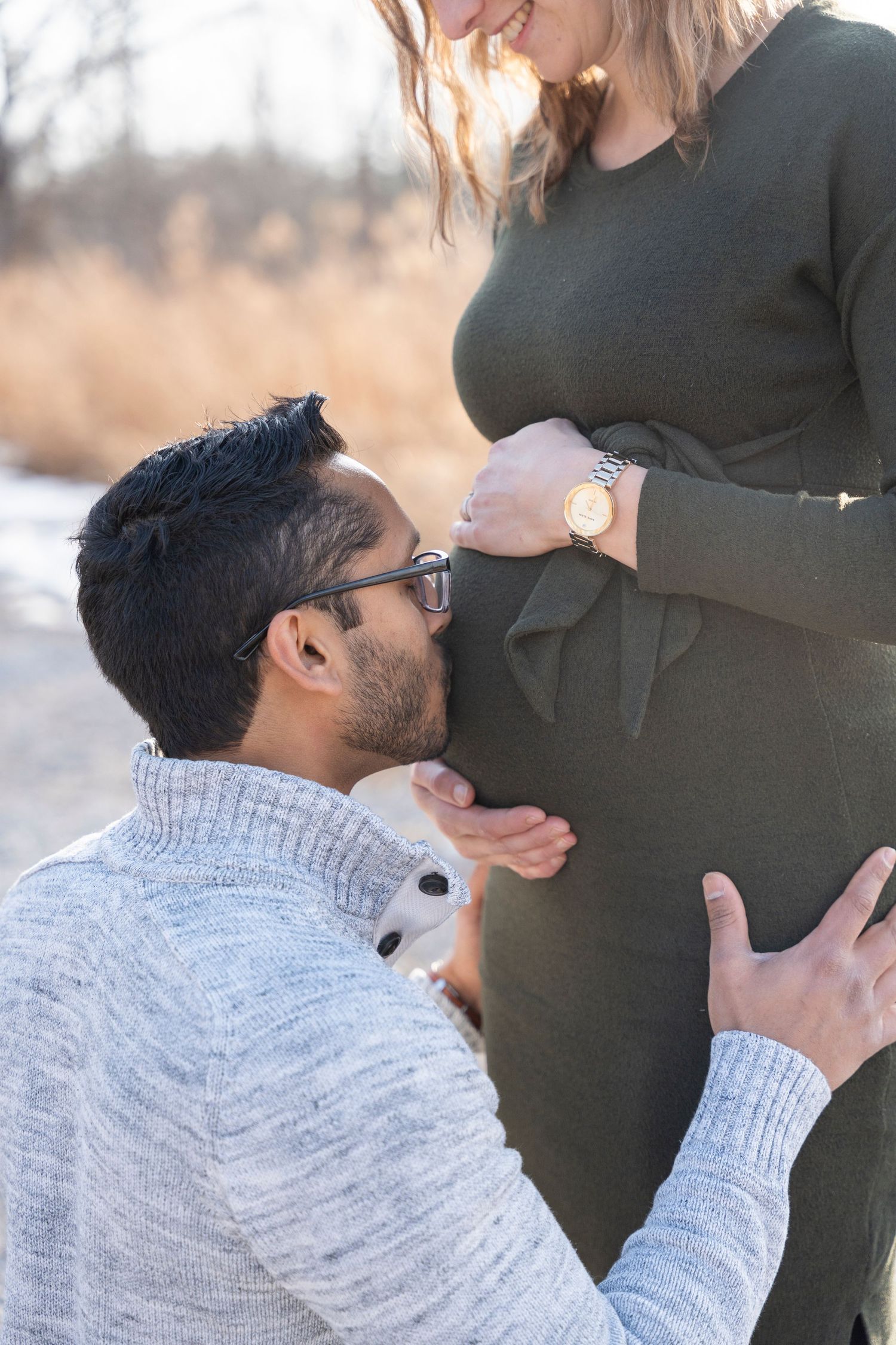 A tender maternity photoshoot moment outdoors with a couple embracing the pregnant belly against a natural backdrop.