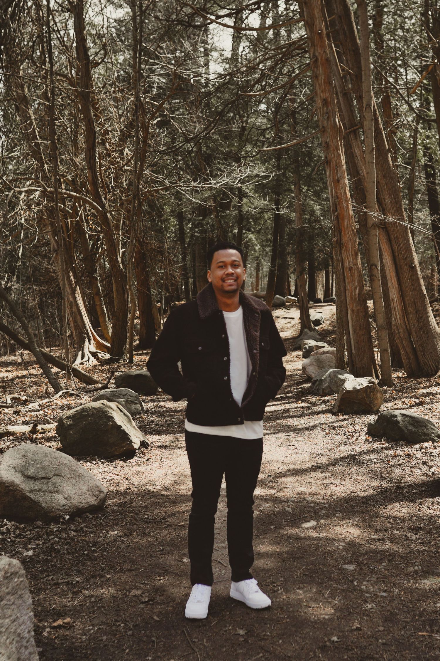 Person in casual wear stands on a forest path surrounded by tall trees.