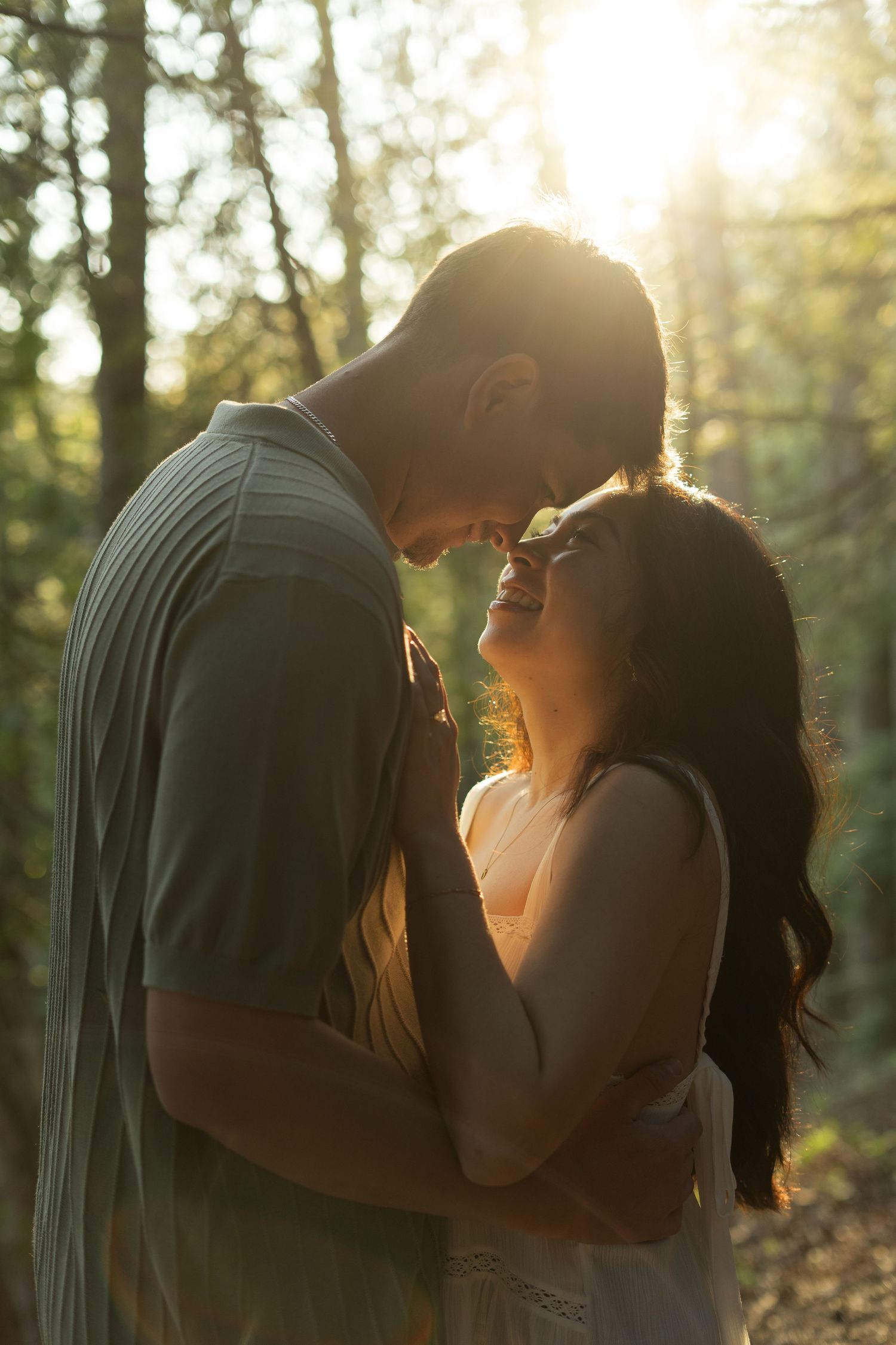 Couple sharing a tender, smiling moment with foreheads touching in a sunlit forest, golden sunlight glowing through the trees behind them.